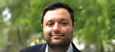 Reyad, a Greek Lebanese American man with a friendly expression, short dark hair and a beard, wearing suit jacket. Background shows blurred green foliage.