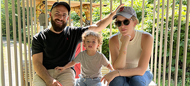 Steve, on the left wearing a dark cap and dark shirt with his female partner wearing sunglasses and a cap and their child outside on a playground