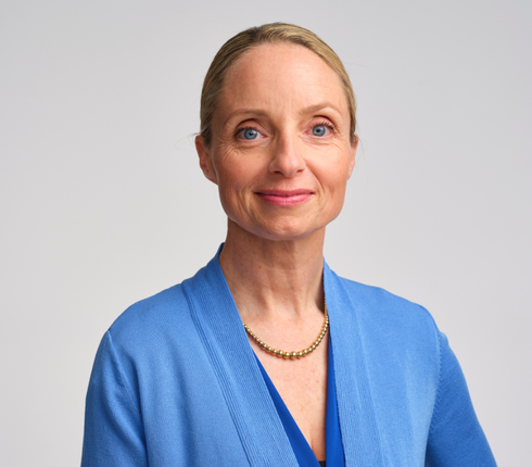 Anne, a white American woman with blonde hair pulled back, wearing a blue blazer and gold necklace, smiling warmly at the camera against a light gray background.