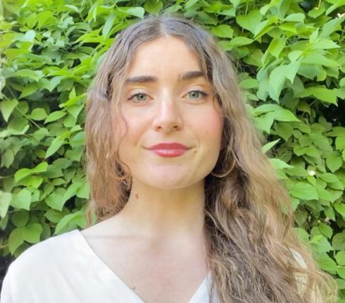 Carlota, long brown wavy hair and brown eyes, wearing a white v-neck shirt.