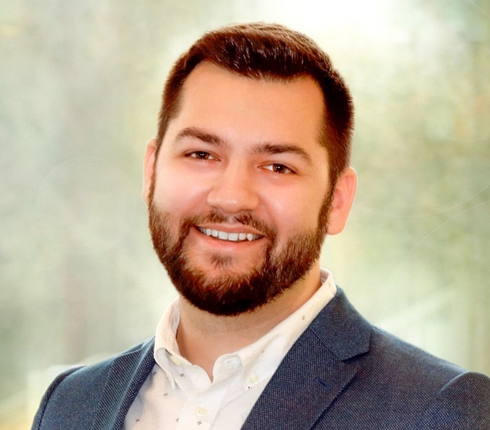 Jake in a professional portrait. Short dark hair and beard, wearing a navy blazer and white shirt against a soft blurred light green background.