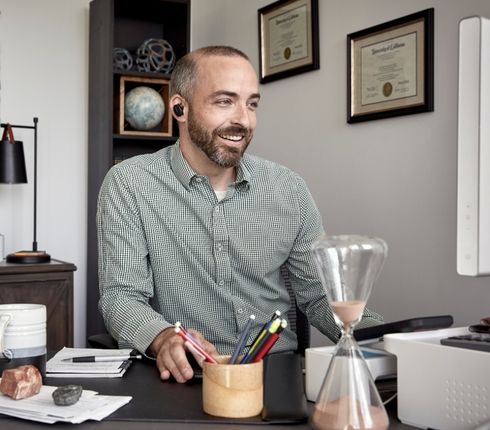 Jared, a white American Parexel employee, works from home. Engrossed in his work, he wears a plaid shirt, has a beard and short hair, and wears headphones while looking at the screen.