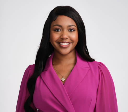 Alexis, a black American woman wearing a pink dress suit and gold heart necklace against a light gray background. She is smiling and looking at the camera.