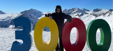 Brando in winter gear standing behind large colorful '3000' sculpture on a snowy mountain plateau with bright blue sky and surrounding alpine peaks in the background.