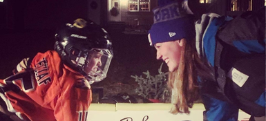 Krista, a woman with long straight hair, wearing winter gear, facing her child who is wearing ice hockey gear, they are smiling at each other.