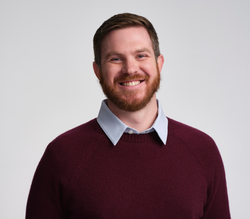 Matt, a white American man, smiling at the camera. He has brown hair and a beard wearing a burgundy sweater over a light blue collared shirt against a neutral gray background.