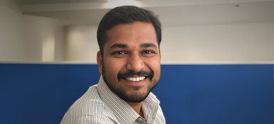 Sameer, a man wearing a light striped shirt seated in an indoor office space with blue partition panels and neutral walls in the background.