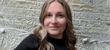 Terra, a woman with long wavy light hair wearing a dark top, standing in front of a textured stone wall with raised patterns and layered designs.