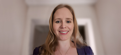 Helen, a woman with long light hair stands indoors in a softly lit hallway, wearing a dark top and a necklace. The background shows neutral-colored walls and doorways slightly out of focus.