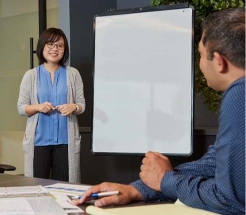 Sammy, a Taiwanese Senior Project Leader, stands in a conference room presenting to another person at a table. She has short dark hair, wears a blue shirt, grey cardigan, and holds a pen.