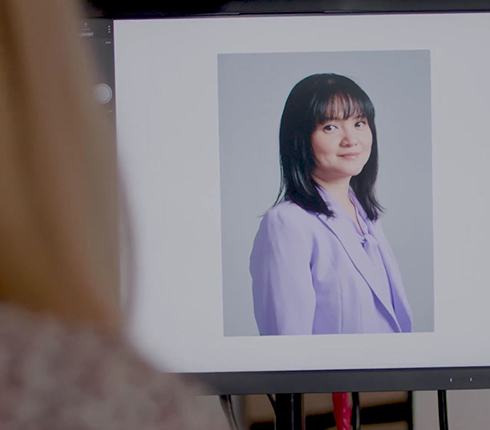 Jo, a Chinese American woman, at the campaign photo shoot. She's wearing a stylish purple jacket and straight long dark hair with fringes. Her picture is being viewed on a screen by a person we see from the back.