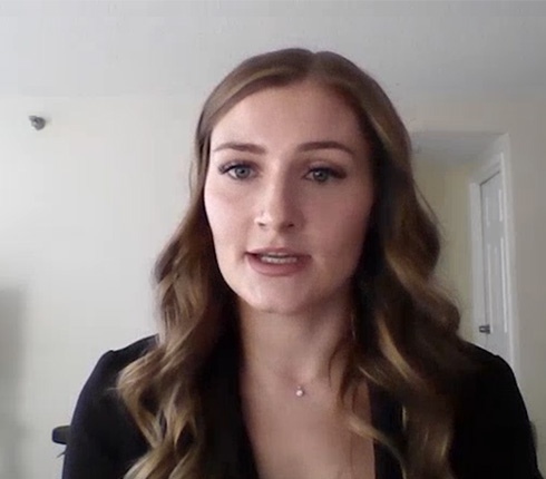 Kylie speaking to the camera. She has long wavy brown hair, wears black blazer and delicate necklace. Her expression is attentive expression. The background indicates a home office with white walls.