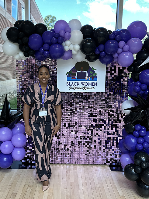 Alexis at the Black Women in Clinical Research conference standing in front of a conference display with purple balloons and a shimmering purple background.