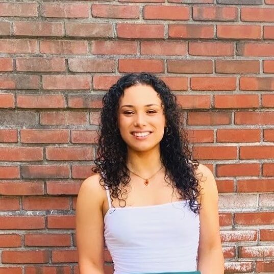 Olivia, a female American, with Pacific Islander and White heritage, is standing in front of a brick wall, she has dark long curly hair and wears a white top.