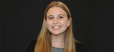 Bella, with straight blonde hair, wearing a black suit jacket and grey shirt is smiling in-front of a black background