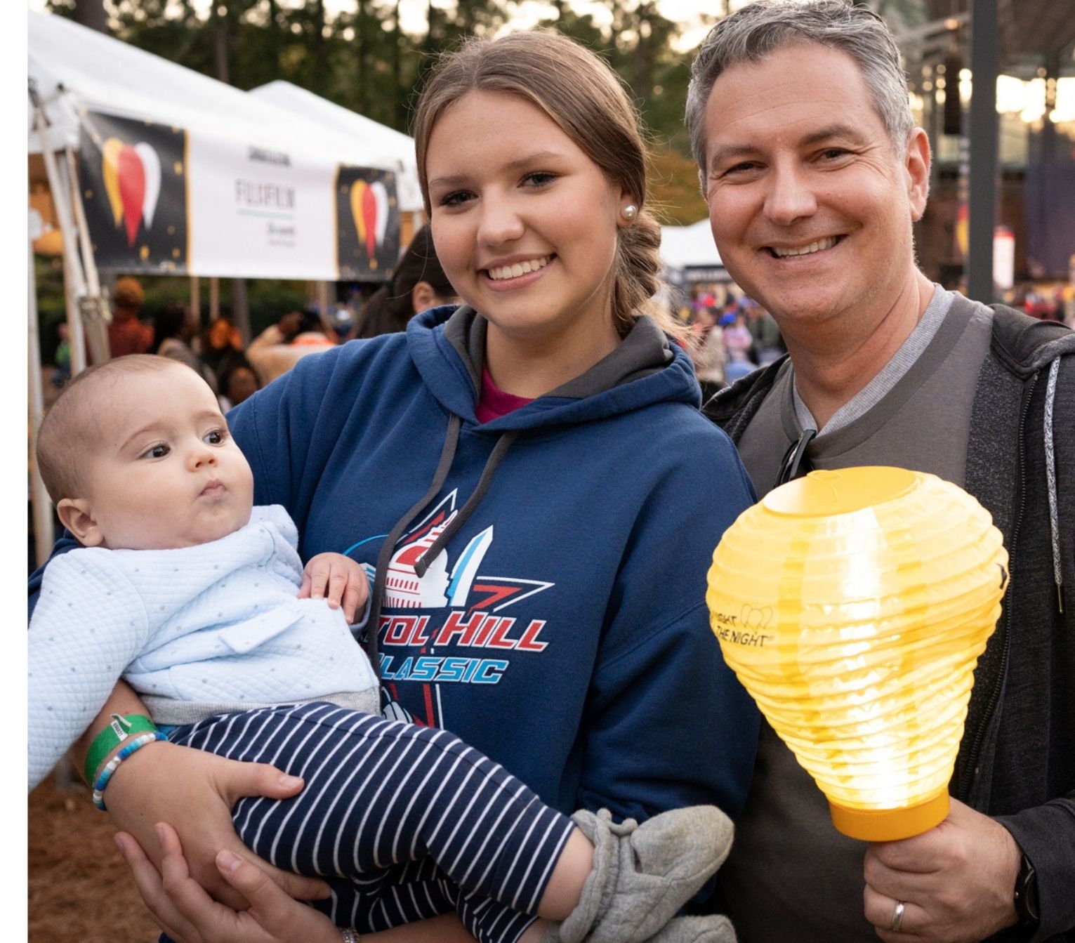 Chris with his daughter. Chris wears a grey sweatshirt and holds a lamp. His daughter wears a blue sweatshirt and is holding a baby.