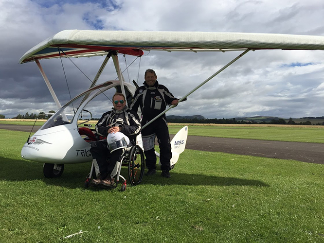 David wearing a dark flying suit with white stripes, and a flight helmet resting on his knee in front o a microlite aircraft with co-pilot similarly dressed stood behind under the wing of the aircraft., sitting in a black and red wheelchair.