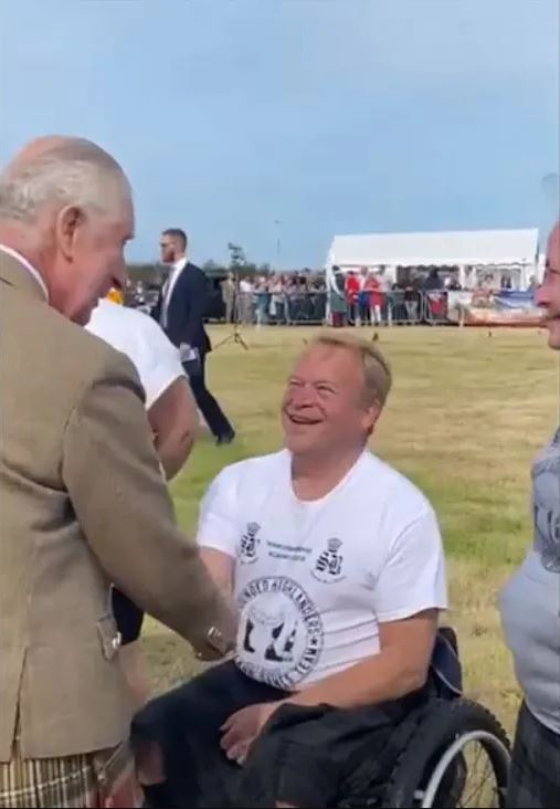 David wearing a white sports shirt in a black and red wheelchair in a field at a sports event shaking hands with King Charles III of the United Kingdom.