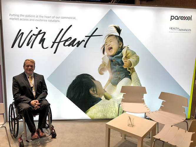 David wearing a grey suit, white shirt, and gold tie, sitting in a black and red wheelchair on a conference stand which has a table and chairs, which displays in the back a highlighted image of the company Parexel and logo ‘With Heart’.