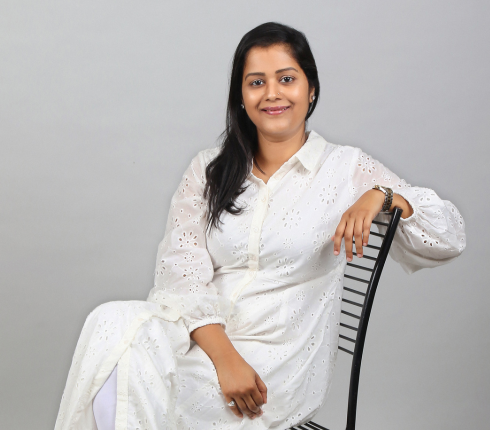 Iram Qayum, An Indian Women, long dark hair, white dress, sitting sideways on a chair with one arm on the back of the chair.