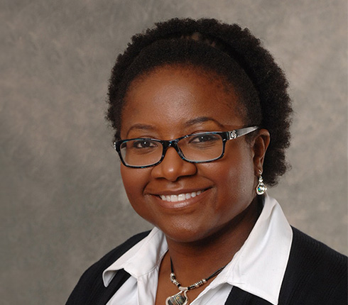 Mwango, a black American woman, she has dark hair tied to the back, wears black glasses. a white shirt and black jacket.