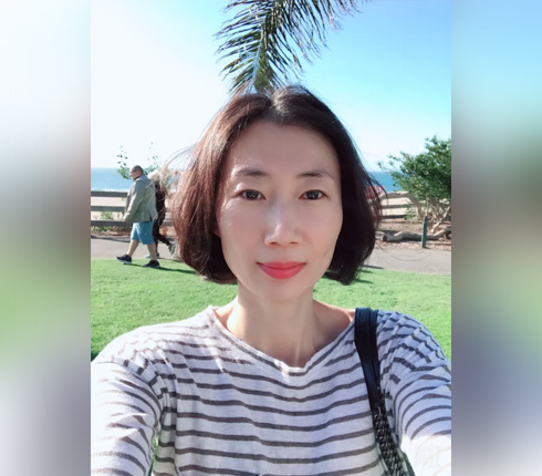 Inhye, a female Korean, with chin long dark hair, red lipstick and a striped shirt taking a selfie with a beach walk in the background.