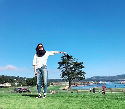 Inhye, a female Korean, full body image stnadng in a landscape, she is holding up a hand marking the hight of a tree, which is further in the back. It creates the illusion that she is taller than the tree.