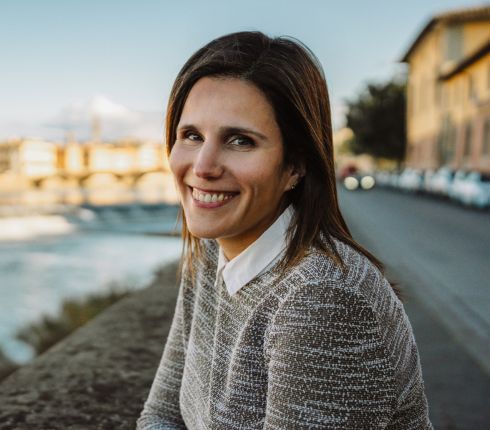 Irene, brown haired woman wearing a grey sweater, smiling while sitting on the bank for a river