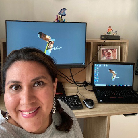 Itzel Pacheco, Parexel Senior Clinical Data Analyst, with dark brown hair pulled into a ponytail wearing a grey t-shirt, sitting in front of two computer screens on a desk.