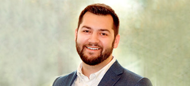 Jake, a white male, with brown hair and beard, is wearing a black suit jacket with a white button up.