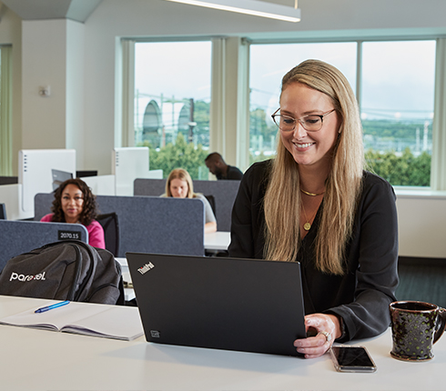 Jessi, a white American Finance professional, is working at a common table in an office. She is smiling at her laptop screen. She has long blond hair and wears a dark shirt.