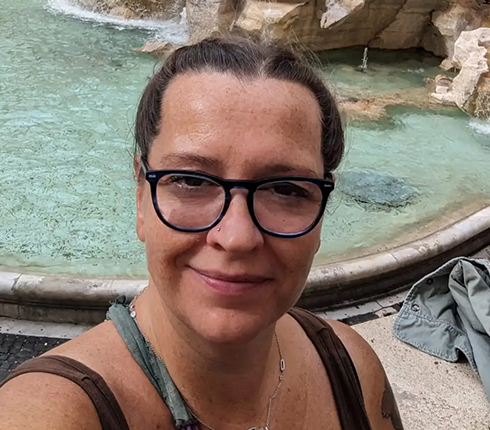 Julie, a female white German, wearing glasses and braided hair, standing in front of a fountain taking a selfie.