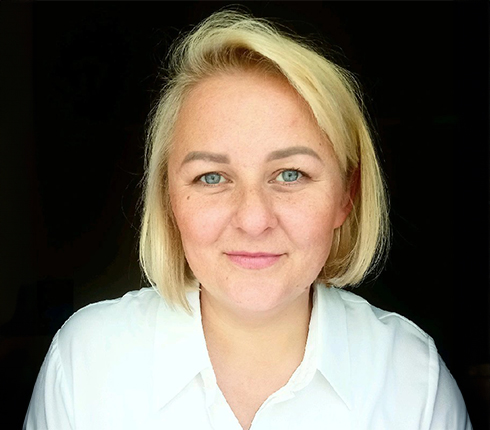 Laura, a Lithuanian woman, with shoulder-length blonde hair, wearing a white shirt against a dark background. She has a kind smile and looks straight at the camera.