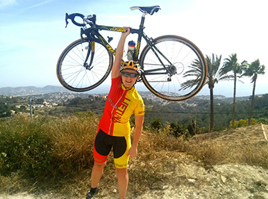 Laura holding up her bike into the air with one hand, she is dressed in a biking outfit and in the background is a dry landscape with palm trees.