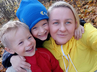 Laura with here two kids, they are outside in a forest with fallen leaves in the background. They all look at the camera and smile.