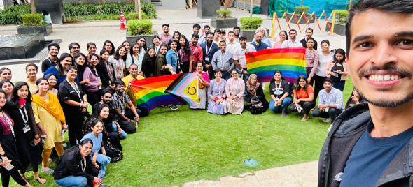 Mayurchandra taking a selfie with a big group of colleagues in the background. The colleagues hold up rainbow-colored Pride Flags.