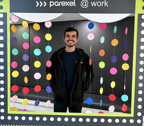 Mayurchandra, an Indian man, in a large picture frame with the text "Parexel @ work" and colorful garlands surrounding him. He has dark short hair and wears a black sweater.