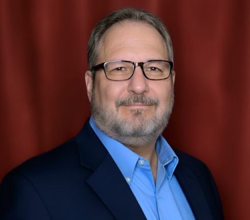 Mitch Cojocariu, a Caucasian man wearing glasses, dressed in a light blue button up shirt and dark suit jacket, smiling