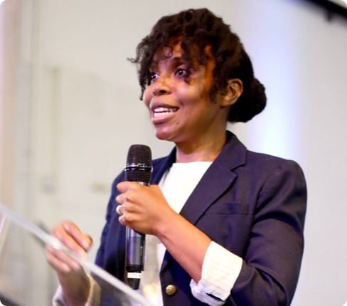 Xoli, a Black English female, giving a talk with a microphone in her hand. She has curly dark hair tied behind in a queue with curly bangs and wears a white shirt with a blue jacket.