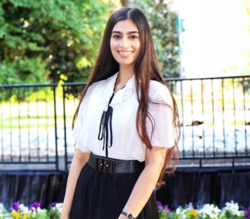 Anooshay, a young woman with long dark hair, smiling at the camera. She wears a white blouse with black tie detail and black skirt. Background shows metal railing, greenery, and flowers in a sunny setting.