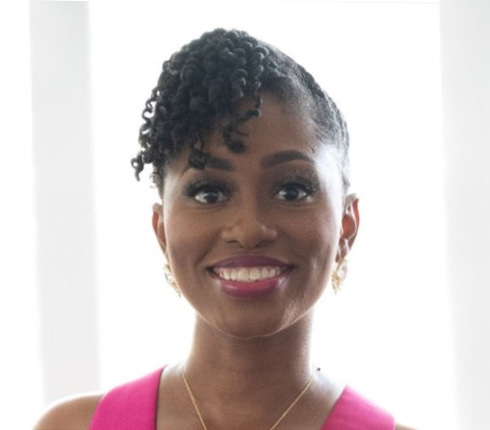A black American lady smiling towards the camera. She wears her hair up, has golden earrings and a golden necklace. She wears a fuscia pink sleeveless top.