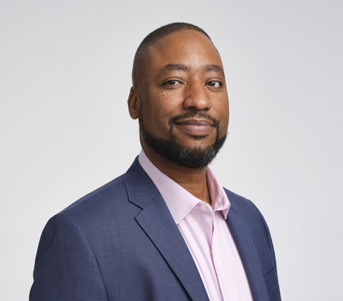 Moe, a Black-American man, wearing short hair and a beard, a blue jacket and a light pink shirt.