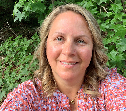 Angela, a female white American, with dark blond curly hair. She wears a flower-patterned shirt and stands in front of leaves.