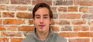 Sean, a male American, dark blond hair and a grey sweater, in front of a brick wall.