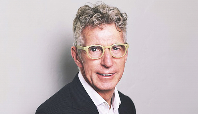 Gwyn Bebb, a white Welsh man, smiling at the camera. He is wearing a black jacket, white shirt, light yellow glasses and grey curly hair.