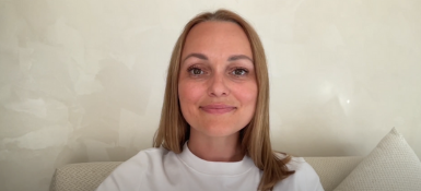 Marina, a young woman with shoulder-length blonde hair smiles at the camera. She is wearing a white sweater and sitting on a light-colored couch or chair. The background appears to be a neutral-toned wall. Her expression is warm and friendly.