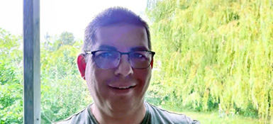 Alan, a smiling middle-aged man with glasses and short gray hair looks at the camera. He's wearing a light-colored shirt. Behind him is a lush green background with a willow tree.