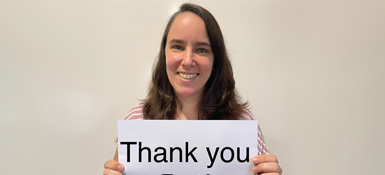 Francisca, a woman with long dark hair holds a white sign that says "Thank you". She's wearing a light-colored top and is standing against a plain, light background. Her expression is warm and appreciative.