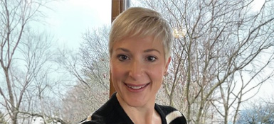 Kris, a woman with short blonde hair looks at the camera. She's smiling at the camera, wearing a black coat with white trim. Behind her are bare trees on a bright and clear winter day.