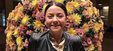 Norah, a woman with dark hair stands in front of a vibrant floral display smiling. She's wearing a black jacket over a light top. The colorful flowers behind her include yellow, pink, and orange blooms, creating a festive backdrop.